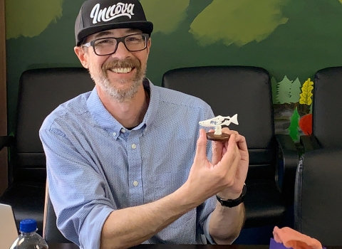 A smiling, bearded man in a ballcap holds up a small carved salmon