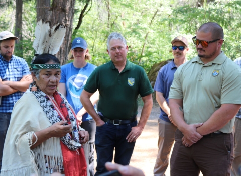 Several people stand in a circle around a woman wearing traditional Tribal garments