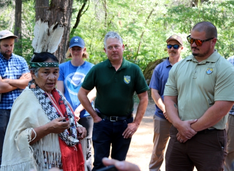 A woman in traditional Native American clothing speaks with several people wearing federal- or state-agency shirts and hats