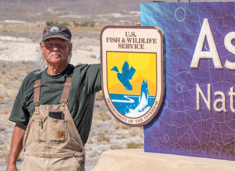 A man with a white mustache wearing overalls and a U.S. Fish and Wildlife Service volunteer cap standing next to a sign with the U.S. Fish and Wildlife Service logo on it