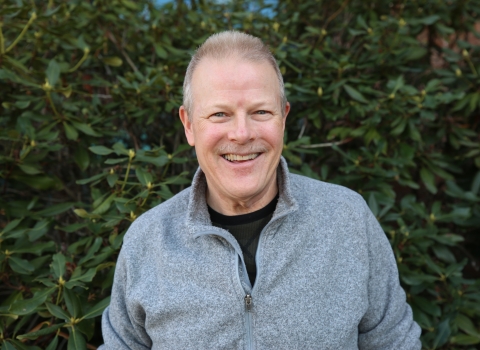 Headshot of a white man in a grey pullover against a leafy backdrop