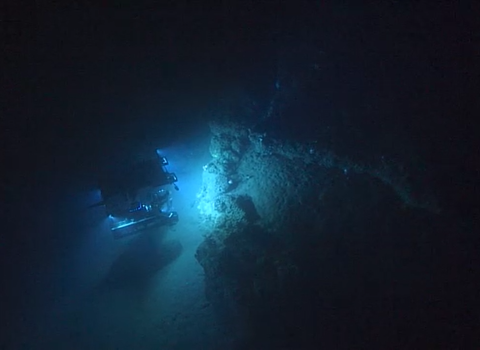 a submarine vessel shines lights on a rocky surface deep underwater. Darkness surrounds the vehicle