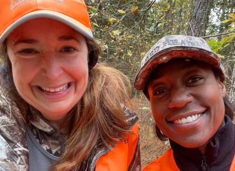 Two women wearing orange and camo, smile together in the woods