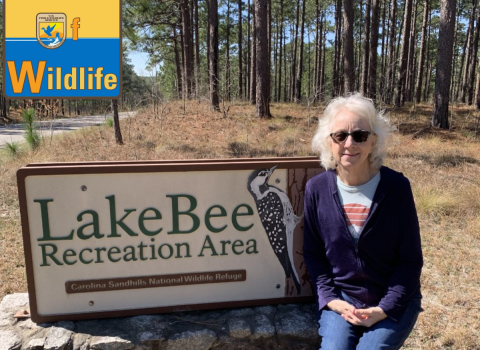 Deb Adams sits on the welcome sign at Lake Bee Recreation area at Carolina Sandhills National Wildlife Refuge. The sign has a woodpecker carved into it. Deb is wearing sunglasses and smiling. The WoW: Woman of Wildlife logo is in the upper left corner. 