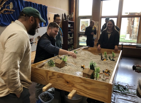 People standing around a table filled with gravel, miniature trees, and miniature sheds