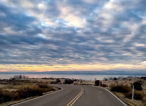 A road in the foreground with a lake and colorful sky with scattered clouds in the background