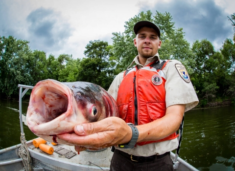 A person wearing a life jacket and standing in a boat, holding up a large fish with open fish mouth facing the camera. 