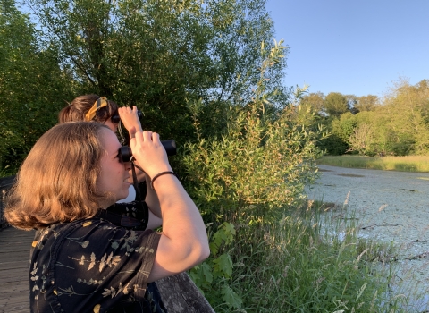 Two people stand at a boardwalk rail with binoculars to the eyes, overlooking a wetland, warm evening light shining from behind them on a clear blue-sky day.