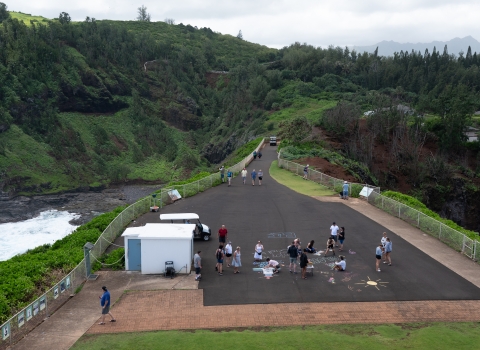 Visitors on a large black walkway at top a bluff are walking and making chalk art. The walk way is surrounded by lush green vegetation and an ocean cove can be seen on the left side. 