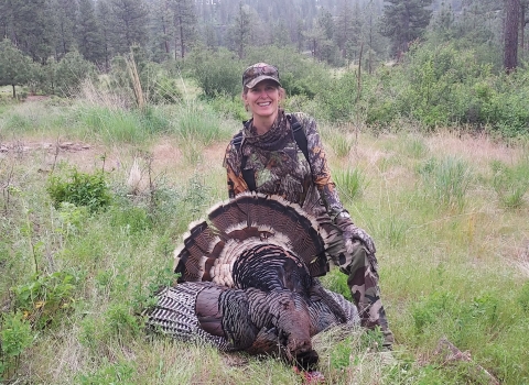 A woman wearing camouflage kneels beneath a turkey she harvested