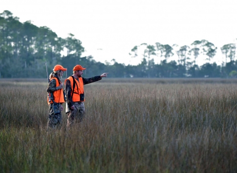 A man and a woman dressed in bright orange safety gear walk across a saltmarsh during a deer hunt