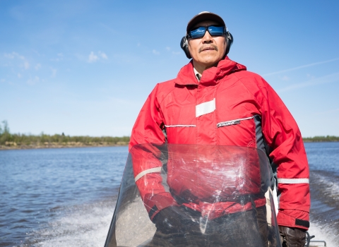 A man in an orange float coat drives a small skiff on a river