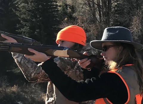 Two women in hunting gear side by side holding long guns side in shooting position