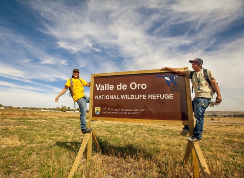 Two smiling young adults from the local community hang off an entrance sign at Valle de Oro National Wildlife Refuge in New Mexico, established in 2012.