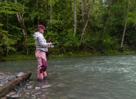 Fisheries intern, Hannah Ferwerda, wading in water and reeling a fishing rod as she samples river for fish.