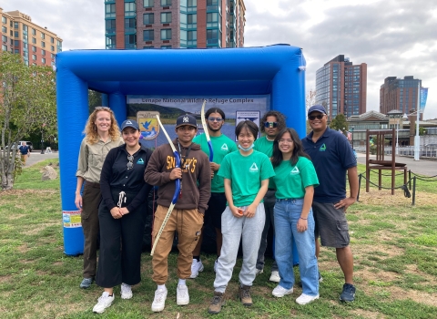 8 people stand in front of a large inflated square object. Some of the people hold archery bows. The group is smiling. It's a cloudy day and there are urban buildings in the background.