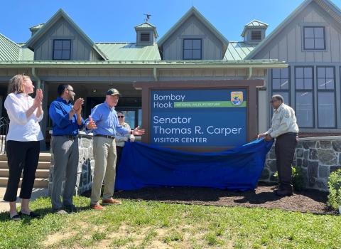 a group of people stand at a large visitor center facility. One of the people pulls off a blue fabric to unveil a sign that says "Bombay Hook Senator Thomas R. Carper Visitor Center". People are clapping in celebration of the event.