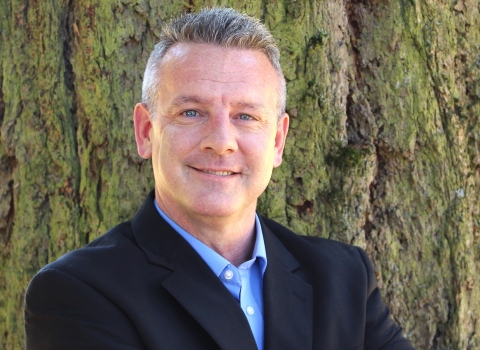 Person smiles for photo wearing dark suit jacket, standing in front of tree.