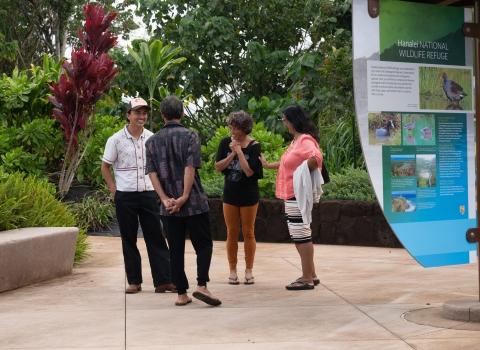 A group of four people stand in a paved walkway next to a sign about endangered wildlife. They are surrounded by lush greenery. Two of the people have huge smiles on their faces. They appear to be chatting. 