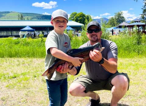 A young boy and his dad show off a caught large trout in front of the fishing pond