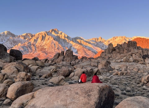 Two people sit on a large rock in the foreground of the picture, with their backs facing the camera as they stare off toward sunrise on the Eastern Sierrva Nevada.