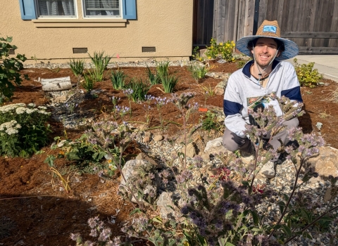 a man wearing a wide brimmed hat squats among plants in a front yard.