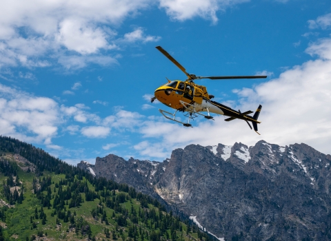 A small yellow helicopter with a crew member looking out soars above rocky mountains in Wyoming.