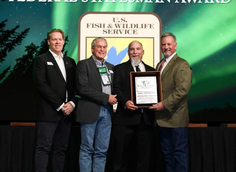 Four people stand onstage and in front of a screen that has the USFWS logo with "Federal Statesman Award." Three men hold an award plaque.