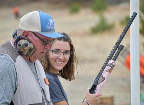 Youth participant in shooting sports holding pink firearm stands next to mentor. 