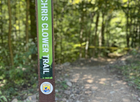 An image of a trail marker that reads "Chris Clower Trail." In the background is a trail that leads through the forest.