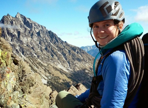 A woman wearing a backpack, rope, and climbing helmet sits high up with a mountain view in the distance