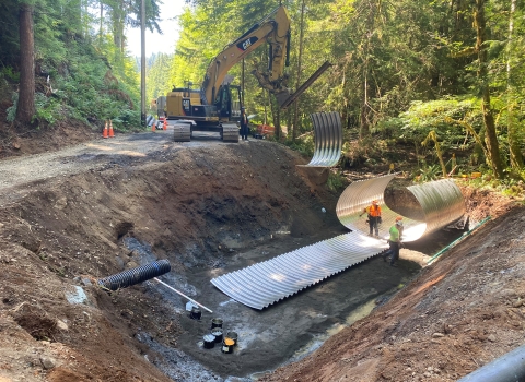 A construction site in the forest with a deep dirt pit with corrugated sheeting and workers wearing safety vests and hard hats. Heavy equipment and trees in the background.