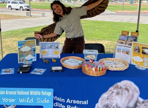 A person smiling and wearing brown wings while standing behind a blue table full of trinkets and educational materials.