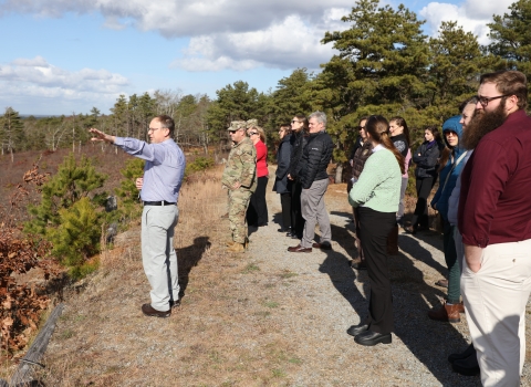 Group of people, some in military camouflage uniforms, outdoors looking off towards the left. A man in front has his arm extended as if he is showing something at a distance