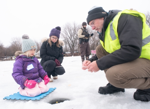 A man in a bright yellow vest assists a young ice angler and her guardian with setting up a fishing rod 