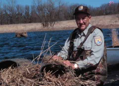 Biologist wearing waders and sitting in a boat on the water fixing a grass/straw hen house that sits on top of the water.
