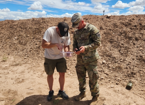 a fish and wildlife service biologist and military personnel dressed in fatigues looks down at a container with a lizard