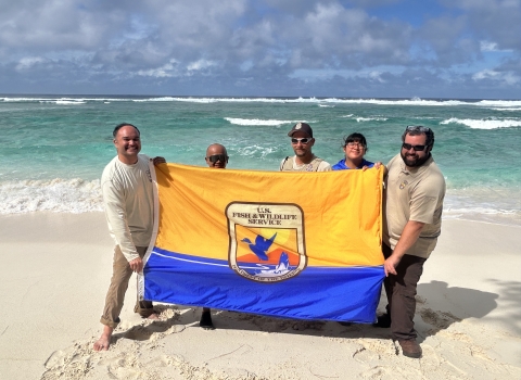 5 people on beach with ocean behind them hold FWS flag