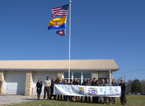 People hold FWS A250 banner at base of flagpole with US flag, FWS flag, fish pennant flying