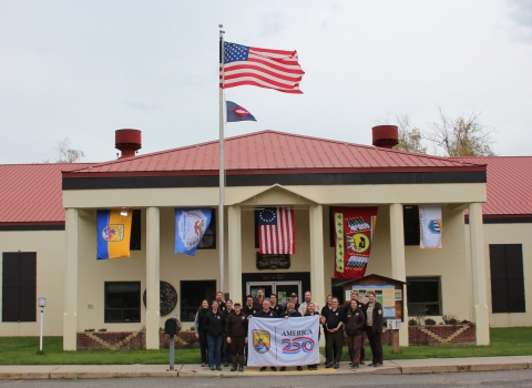A group of people in brown holding a white “America 250” flag stand in front of a tan building with a red roof. Flags from USFWS, the Confederated Tribes and Bands of the Yakama Nation, the Betsy Ross flag, the Confederated Tribes of the Colville Reservation, and the Bureau of Reclamation flag fly behind. US flag and fish pennant fly above