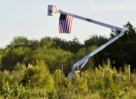 An American flag flies on a lift over dense green shrubs and trees.