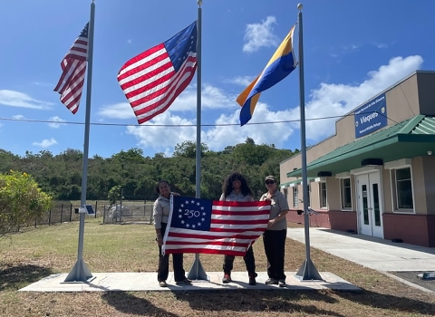 Three people hold a Freedom 250 flag in front of a building, with U.S. and FWS flags flying on poles